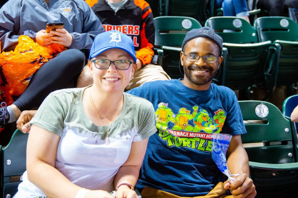 A nice young couple sitting in their seats smiling for the camera. The girl has the gvsu night tigers hat on and the guy is wearing an awesome teenage mutant ninja turtles shirt